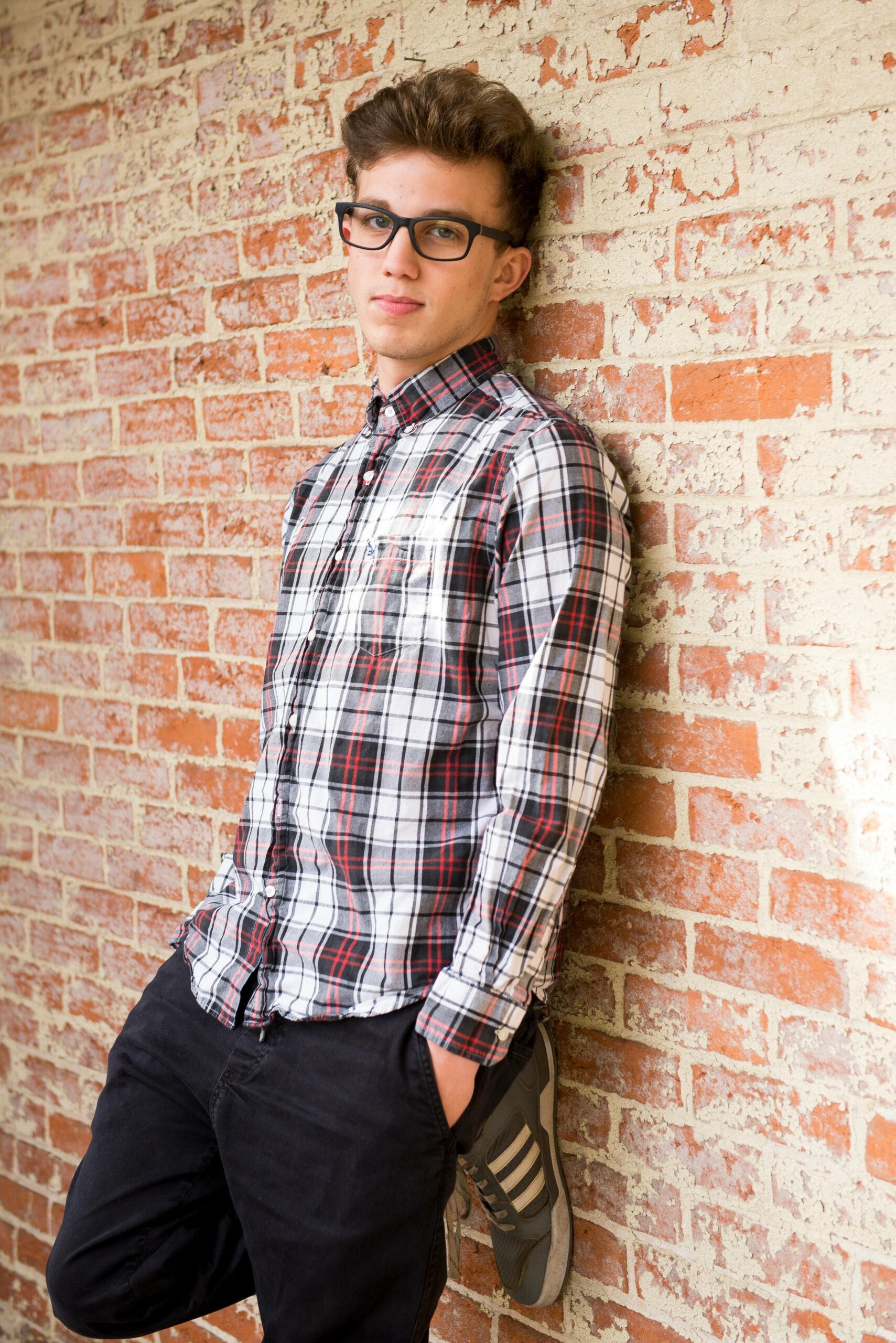 Stylish young man in casual plaid shirt and eyeglasses leaning against a brick wall.